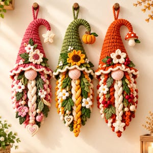 Puede incluir: Tres decoraciones de pared de gnomos de ganchillo con sombreros puntiagudos. El gnomo de la izquierda es rosa con flores, el central es verde con un girasol y una calabaza, y el de la derecha es naranja con una seta y bayas. Barba trenzada.