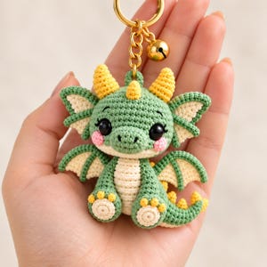 May include: A crochet dragon keychain featuring a green body, cream-colored belly, and yellow horns and spikes. The dragon has large black eyes, pink cheeks, and small wings. It is attached to a gold-toned metal ring with a small bell.