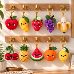 Puede incluir: Diez llaveros de frutas tejidos a ganchillo colgados en dos estantes de madera. La colección incluye una manzana roja, un mango amarillo, una fresa roja, uvas moradas, un plátano pelado, dos cerezas rojas, una piña, una sandía, una naranja y un limón.