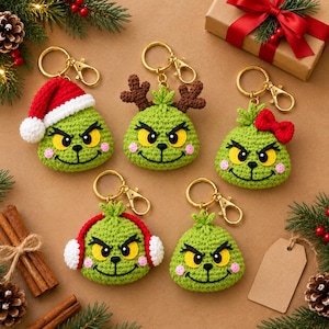 May include: Five crocheted keychains featuring green, round faces with yellow eyes, black eyebrows, and pink cheeks. One wears a red Santa hat, one has brown antlers, one has a red bow, one has red earmuffs, and one is plain.