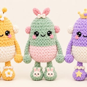 May include: Three crocheted plush figures with rounded bodies, smiling faces, and heart-shaped eyes. The yellow figure has a daisy, the green one has rabbit ears, and the purple one wears a crown. Each has white midsections and unique footwear.