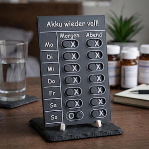 May include: A dark gray rectangular medication tracker board with white text reading Akku wieder voll, Morgen, and Abend. It features rows for days of the week from Mo to So, each with two sliding toggle switches marked with an O and an X symbol.