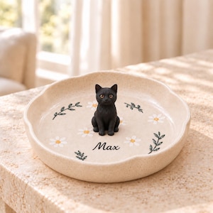 May include: A round, speckled beige ceramic trinket dish with a scalloped edge, featuring a small, three-dimensional black cat figurine in the center. The dish is decorated with white daisy illustrations, green leaf sprigs, and the name Max.