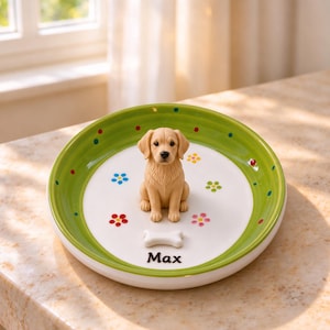 Puede incluir: Un plato redondo de cerámica con borde verde y pequeños puntos de colores. En el centro hay una figura de perro de color marrón claro, un objeto blanco con forma de hueso y el nombre Max impreso en letras negras. La base blanca tiene flores.