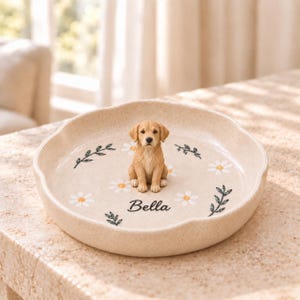 Puede incluir: Un plato decorativo redondo de cerámica beige moteada con una pequeña figura tridimensional de un golden retriever en el centro. El plato está decorado con margaritas blancas pintadas y ramas verdes, con el nombre Bella impreso en negro.