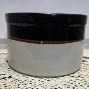May include: A cylindrical ceramic crock featuring a two-tone design with a dark brown glazed upper section and a light beige lower section. The vessel has a smooth, glossy finish on the top rim and a matte texture on the base.