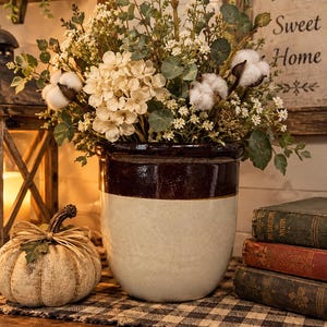 May include: A two-toned ceramic crock with a dark brown glazed upper section and a cream-colored lower body. The crock is filled with an arrangement of white cotton bolls, small white flowers, and green eucalyptus-like foliage stems.