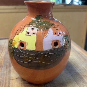 May include: A terracotta vase with a rounded body and a narrow neck. The surface features a hand-painted scene of two small houses, one yellow and one light purple, situated above a dark brown area representing water, with green accents.