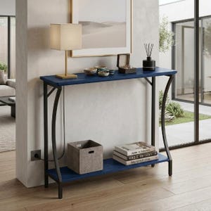 May include: A navy blue console table with a black metal frame, featuring two shelves. The top shelf holds a beige lamp, small bowls, and a black box. The bottom shelf contains a grey storage bin and a stack of three books titled Architecture, Vogue The Gown, and Modern Forms.