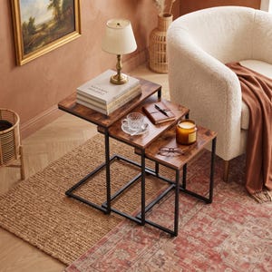 May include: A set of three nesting tables featuring rectangular wooden tops with a dark brown finish and black metal frames. The tables are arranged in a stepped formation, with the largest table supporting a lamp, books, and a small cup.