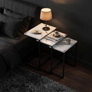 May include: A set of three nesting tables with white faux marble tops and black metal frames. The tables are arranged in a stepped formation, holding a table lamp, a small dish with keys, two stacked books, a ceramic mug, and a small potted succulent.