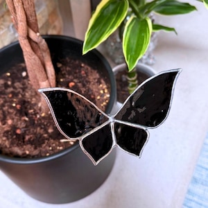 May include: A stained glass butterfly ornament featuring four glossy black wings outlined in a thin silver metal solder. The butterfly is positioned horizontally with a symmetrical wing span, showcasing a smooth, reflective surface and a clean, defined edge.