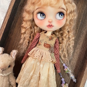 May include: A doll with long wavy blonde hair and large blue eyes wearing a tan dress with a brown polka-dot long-sleeve top underneath. The dress features a basket print on the bodice and a white floral embroidered hem. A small plush toy is nearby.