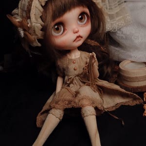 Pode incluir: Uma boneca com grandes olhos castanhos e cabelo castanho, vestindo um vestido bege com acabamento em renda, uma touca a condizer com folhos, meias bege até ao joelho e sapatos castanhos escuros. A boneca segura uma fina corrente dourada.