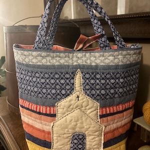 May include: A quilted tote bag featuring horizontal fabric strips in shades of blue, coral, yellow, and white. The front displays a cream-colored fabric applique of a church building with a small cross and a blue patterned door. Two fabric handles.