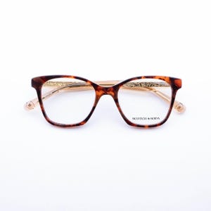 May include: Tortoiseshell-patterned eyeglasses with a brown and amber frame. The temples feature gold-colored metal with an intricate engraved pattern and a small floral detail at the tips. The right lens displays the text SCOTCH & SODA.