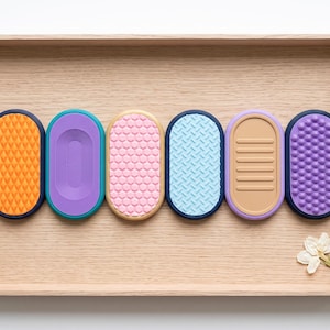 Könnte beinhalten: Sechs ovale sensorische Silikonpads mit unterschiedlichen Texturen und Farben, darunter Orange, Lila, Rosa, Hellblau, Beige und Dunkellila, in einer Reihe angeordnet. Jedes Pad weist ein einzigartiges Oberflächenmuster wie Rauten oder Rillen auf.