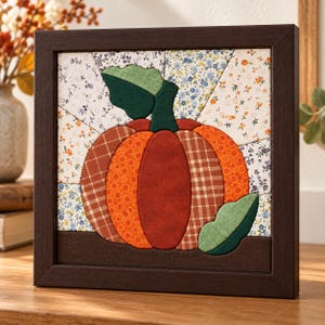 Pode incluir: Uma peça de arte de parede em tecido emoldurada de forma quadrada com uma aplicação central de abóbora feita de vários tecidos estampados em laranja e castanho. A abóbora tem duas folhas de tecido verde sobre um fundo de segmentos de tecido floral.