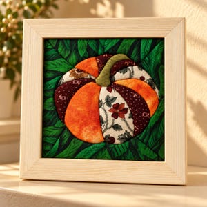 Pode incluir: Uma peça de arte de parede em tecido emoldurada de forma quadrada com um motivo central de abóbora. A abóbora é construída com segmentos de tecido acolchoado em laranja, bordô escuro e creme com padrões florais, rematada com um caule verde sobre folhas.