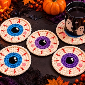 Puede incluir: Un conjunto de cinco posavasos redondos de ganchillo con forma de globo ocular. Cada posavasos presenta una esclerótica de color crema con venas rojas bordadas, una pupila negra central con reflejos blancos y un iris azul o morado con detalles.