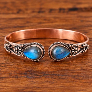May include: A copper cuff bracelet featuring two teardrop-shaped labradorite gemstones with blue iridescence. The metal band is decorated with twisted wire borders, small metal beads, and scrollwork patterns at both open ends.