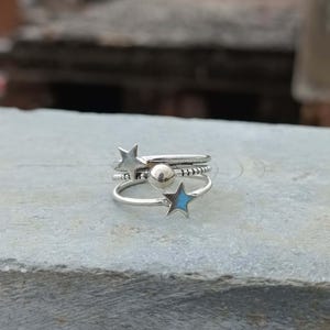 May include: A silver-toned multi-band ring featuring three distinct layers. The top band has a star shape, the middle band includes a small spherical bead, and the bottom band displays a star with a light blue inlay. The metal has a polished finish.