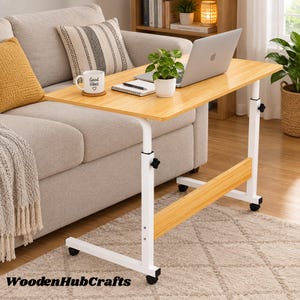 May include: A mobile laptop desk with a light wood-grain rectangular top and a white metal frame. The desk features adjustable height knobs, a lower support beam, and four black caster wheels. A laptop, a notebook, and a mug are on the surface.