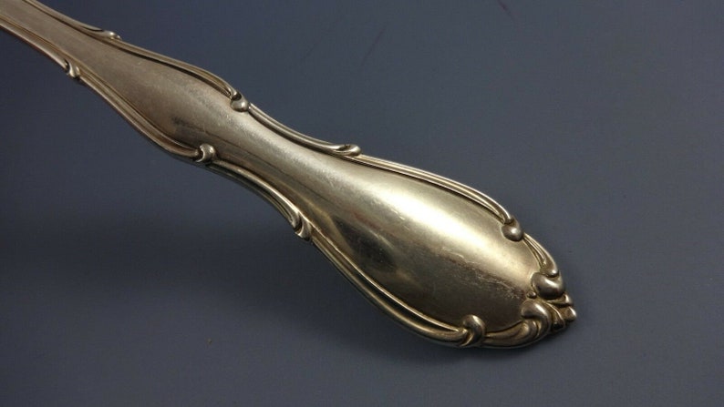 May include: Close-up of the handle of a silver fork with an ornate design. The handle is smooth and shiny.