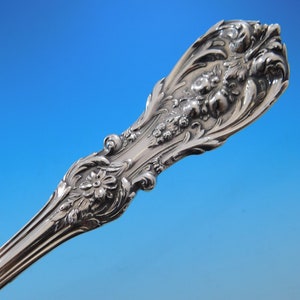 May include: Close-up of a silver utensil handle with ornate floral and scrollwork detailing. The handle features a flower and berry design, with a polished, reflective surface. The background is a gradient of blue.