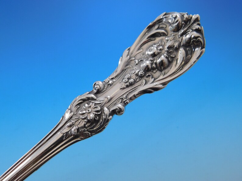 May include: Close-up of a silver utensil handle with ornate floral and scrollwork detailing. The handle features a flower and berry design, with a polished, reflective surface. The background is a gradient of blue.