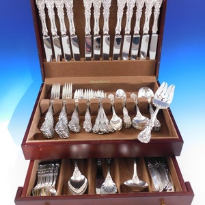 May include: A full set of silver cutlery, including knives, forks, and spoons, is presented in a dark wooden case with multiple drawers. The cutlery features elaborate floral detailing on the handles. The case is lined with brown fabric.