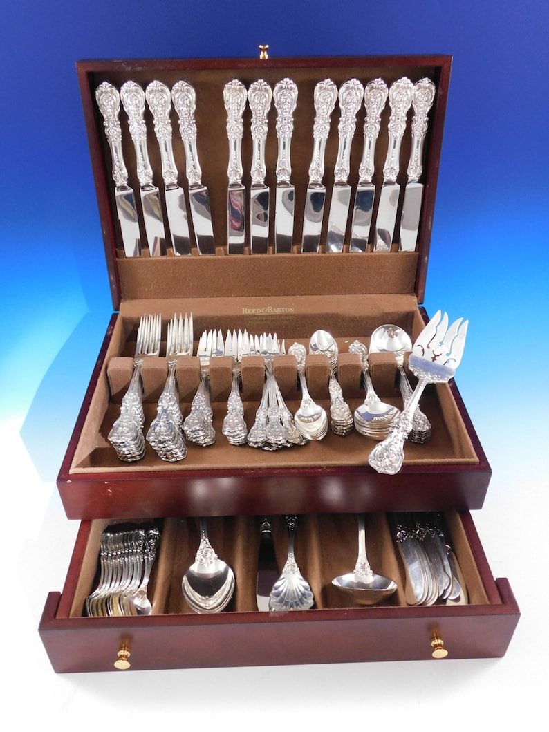 May include: A full set of silver cutlery, including knives, forks, and spoons, is presented in a dark wooden case with multiple drawers. The cutlery features elaborate floral detailing on the handles. The case is lined with brown fabric.