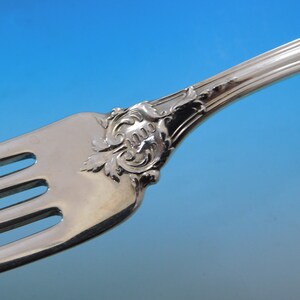 May include: Close-up of a silver fork with ornate detailing. The fork features three tines and a decorative handle with a floral and scroll design. The silverware is set against a gradient blue background.
