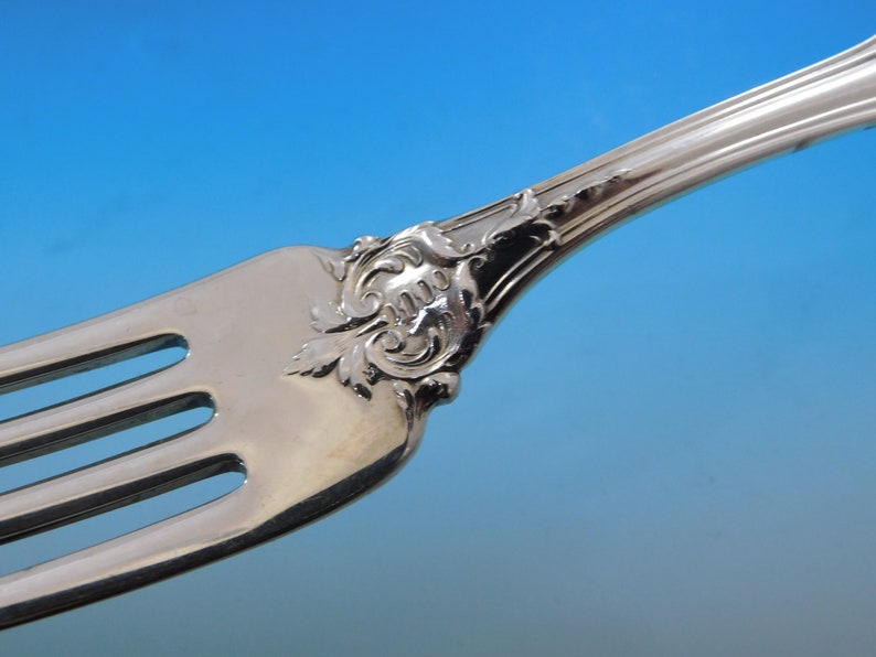 May include: Close-up of a silver fork with ornate detailing. The fork features three tines and a decorative handle with a floral and scroll design. The silverware is set against a gradient blue background.