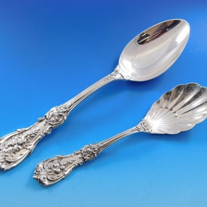 May include: Two ornate silver serving spoons against a blue backdrop. One spoon features a large, oval bowl and a detailed floral handle. The other has a shell-shaped bowl and a matching floral handle.