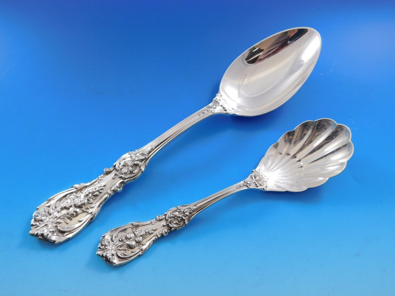 May include: Two ornate silver serving spoons against a blue backdrop. One spoon features a large, oval bowl and a detailed floral handle. The other has a shell-shaped bowl and a matching floral handle.
