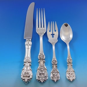 May include: A collection of four decorative silver-coloured cutlery items: a knife, a fork, a serving fork, and a spoon. The handles of each piece are adorned with intricate floral and scroll designs. The knife blade is polished.