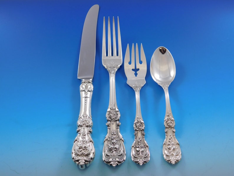 May include: A collection of four decorative silver-coloured cutlery items: a knife, a fork, a serving fork, and a spoon. The handles of each piece are adorned with intricate floral and scroll designs. The knife blade is polished.
