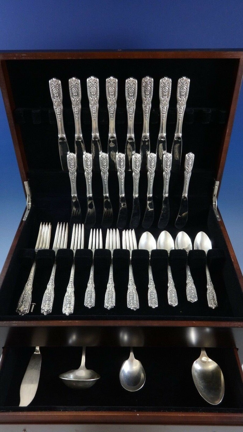 Puede incluir: Una caja de madera con un interior de terciopelo negro que contiene un juego de cubiertos de plata. El juego incluye cuchillos, tenedores, cucharas y piezas para servir. Los cubiertos son ornamentados y presentan un dise&ntilde;o floral.