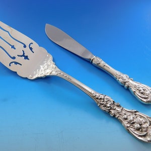 May include: Silver serving fork and butter knife set. The fork has a decorative pierced design and the knife has a rounded blade. Both pieces feature ornate floral and scrollwork detailing on the handles.
