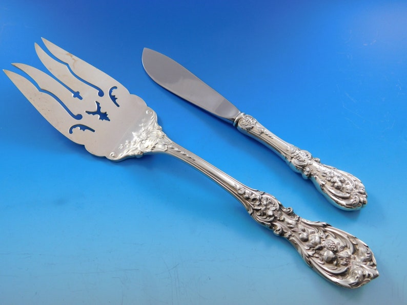 May include: Silver serving fork and butter knife set. The fork has a decorative pierced design and the knife has a rounded blade. Both pieces feature ornate floral and scrollwork detailing on the handles.