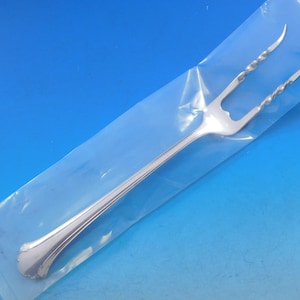 May include: A silver serving fork with a long handle and two tines. The tines have a twisted design. The fork is in a clear plastic bag against a blue background. The fork is likely for serving food.