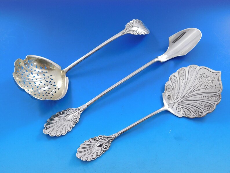 May include: Three silver serving spoons with ornate handles. The spoons have different shapes and designs, including a spoon with a perforated bowl.