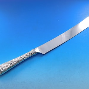 May include: A silver cake knife with a detailed floral design on the handle and a serrated blade.