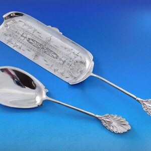 May include: A set of two silver serving spoons with ornate handles. The larger spoon has a decorative, engraved design on the bowl. Both spoons have a polished finish.