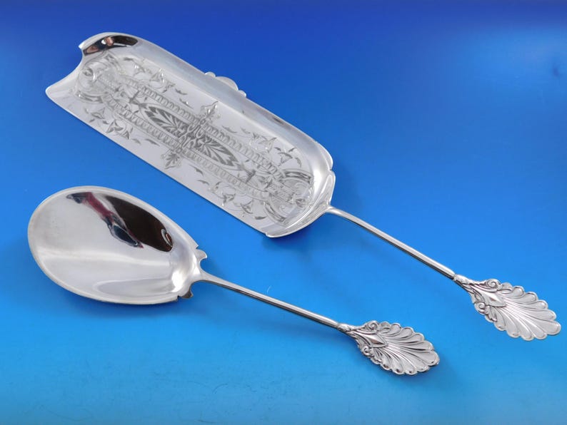May include: A set of two silver serving spoons with ornate handles. The larger spoon has a decorative, engraved design on the bowl. Both spoons have a polished finish.