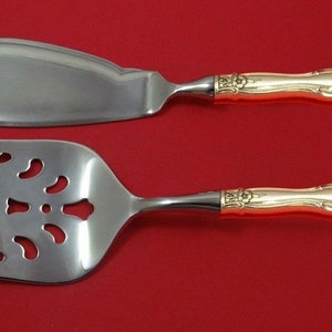 May include: A silver-colored serving fork and cake server set with ornate gold-colored handles. The fork has a slotted design, and the cake server has a curved blade. The set is displayed against a red background.