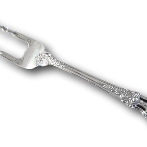 May include: A silver serving fork with a decorative floral pattern on the handle. The two tines have a twisted design. The fork is likely made of stainless steel or silver and is designed for serving food. The fork is on a white background.