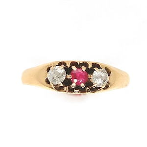 May include: A gold ring featuring a central red gemstone flanked by two clear gemstones. The ring has a classic design with a smooth band and decorative settings around the stones. The gemstones are round-cut and set in a floral-like pattern.