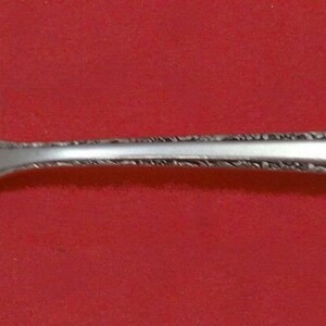May include: A silver serving spoon with a slotted, oval bowl and a decorative handle. The bowl features a stylized floral design. The handle is detailed with an ornate pattern. The spoon is set against a red background.